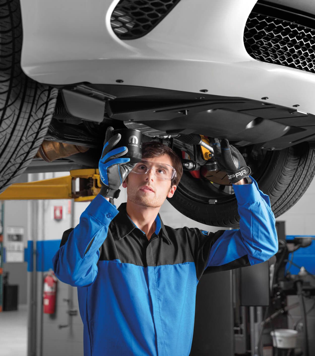Male mechanic in a Mopar uniform replacing an oil filter underneath a white vehicle raised on a shop lift.