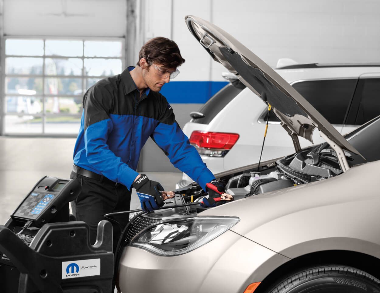 Male Mopar mechanic using diagnostic equipment on a vehicle battery under an open hood in a service center.
