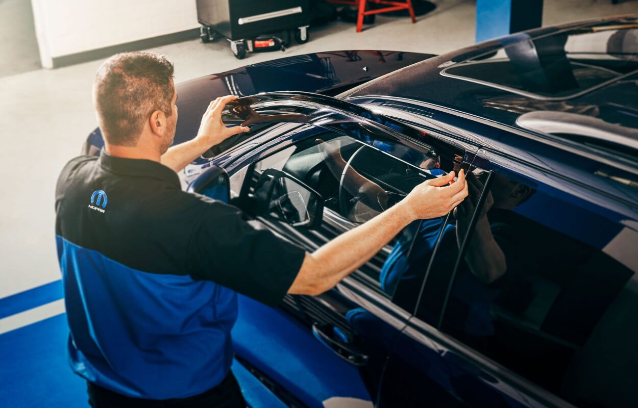 High-angle view of a Mopar technician installing a dark window deflector accessory onto a blue vehicle.