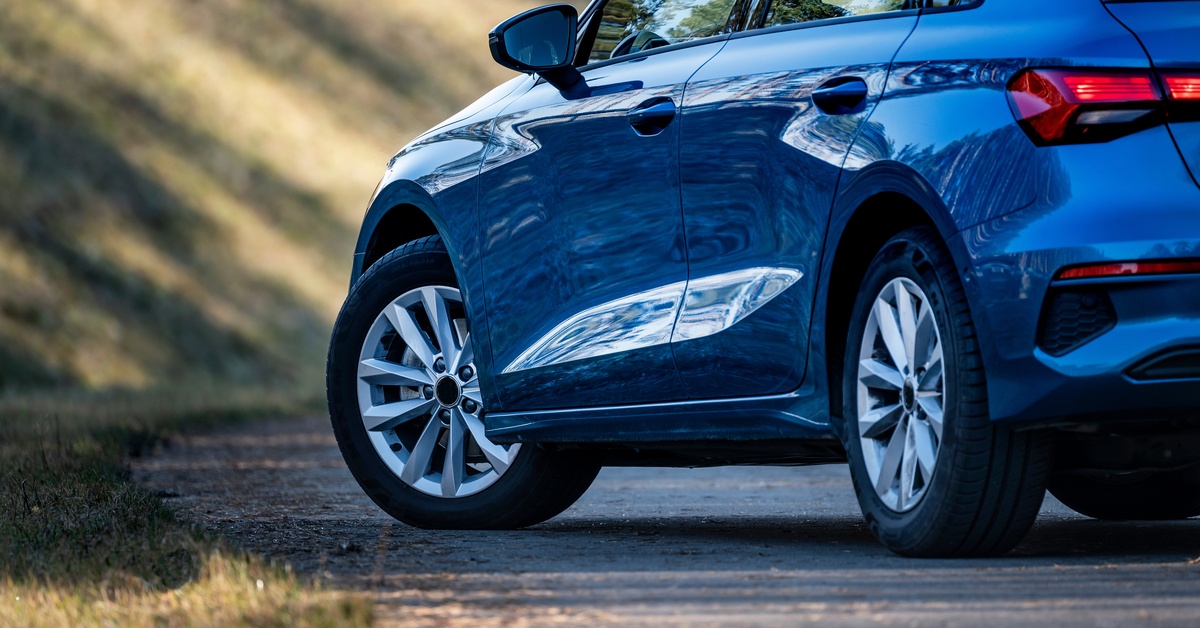 A blue Audi parked on the side of the road. The car is covered in shadow but some sunlight is hitting the grass nearby.