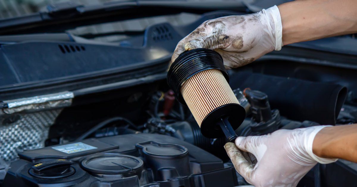 A person wearing white gloves is standing in front of a car with its hood open. The person is holding an oil filter.