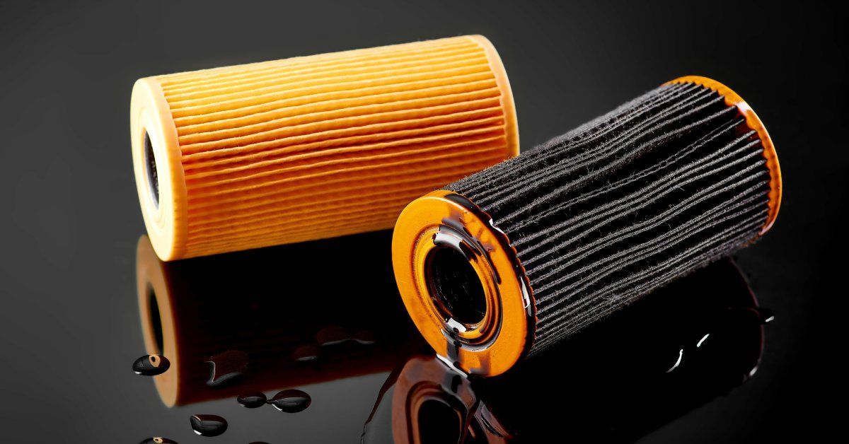 An orange oil filter and a black oil filter lying on a shiny black surface. Several drops of oil are near the filters.
