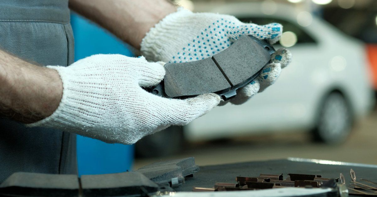 A person wearing white gloves with blue dots stands next to a white car while holding a black brake pad.