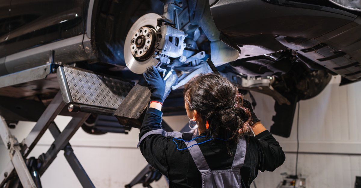 A mechanic wearing black gloves uses a light to inspect a car’s suspension system. The car’s tires have been removed.