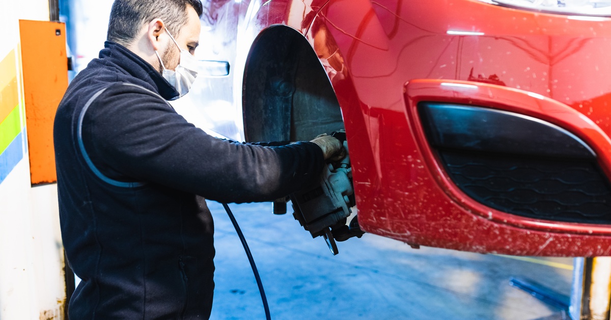 A person wearing a white mask, installing a new brake rotor on a shiny red car. The sun shines on the car.