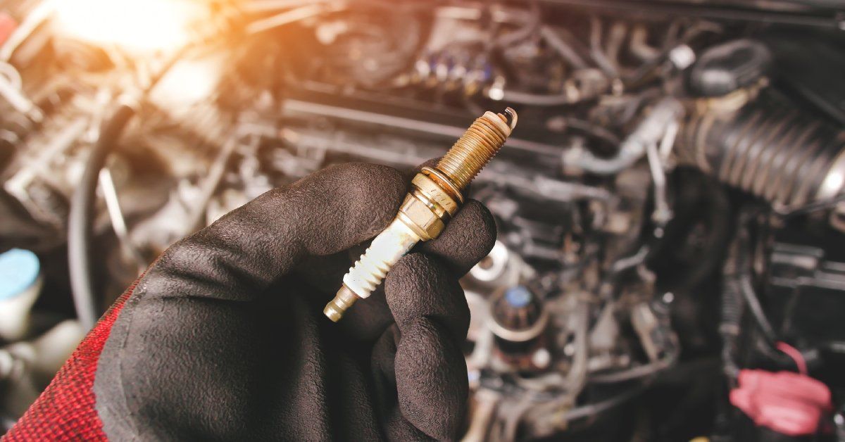 A person wearing thick, red-and-black work gloves holding a small spark plug over the open hood of a car. A person wearing thick, red-and-black work gloves holding a small spark plug over the open hood of a car.