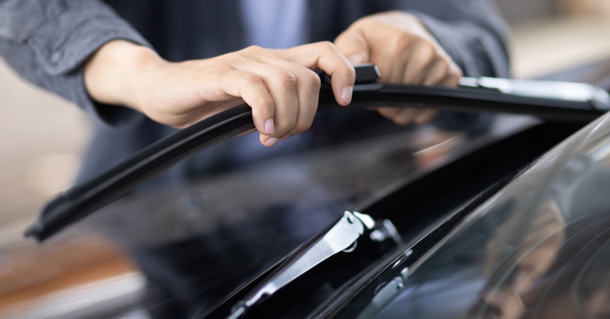 A person standing next to a car is leaning over the hood to install a new black windshield wiper blade.