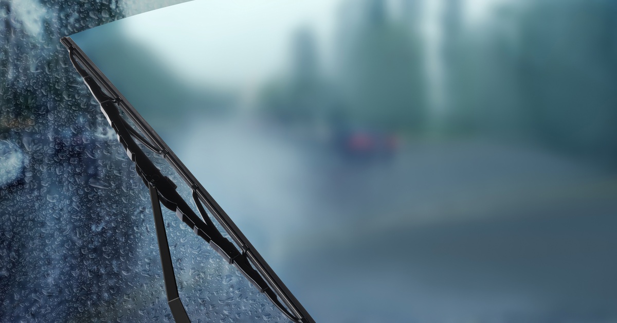 A small, thin, and black windshield wiper blade slides across a car windshield covered in rain drops.