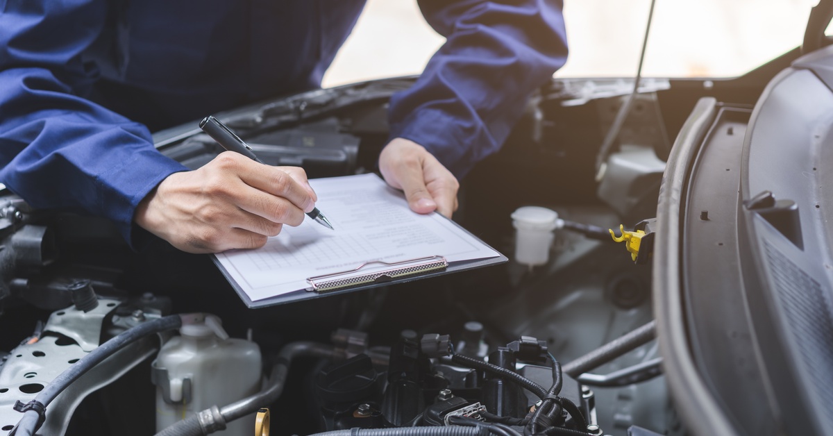 A vehicle with its hood open. A person wearing a blue shirt leans over the hood while holding a pen and a clipboard.