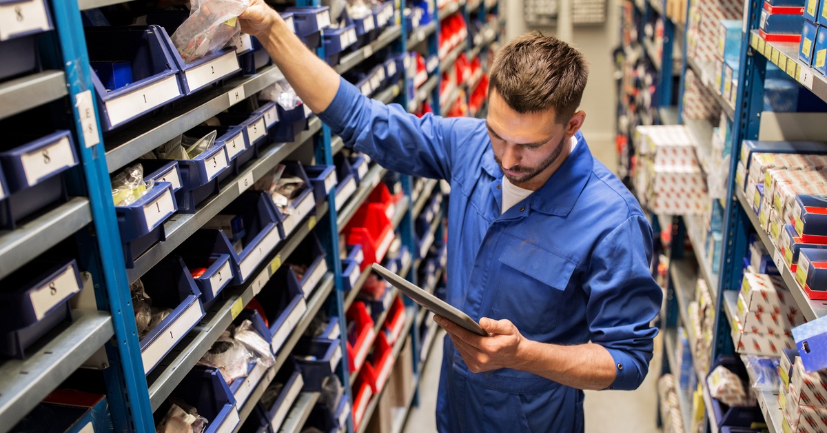 An employee reaches for a product on a store shelf with their right hand while looking at a tablet in their left hand.