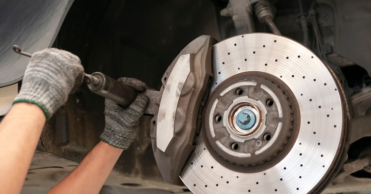 A person wearing gray gloves is installing brake pads on the underside of a vehicle in an automotive shop.