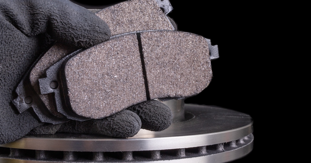 A person wearing black gloves holding two new brake pads in one hand. A metal disk is visible behind the brake pads.
