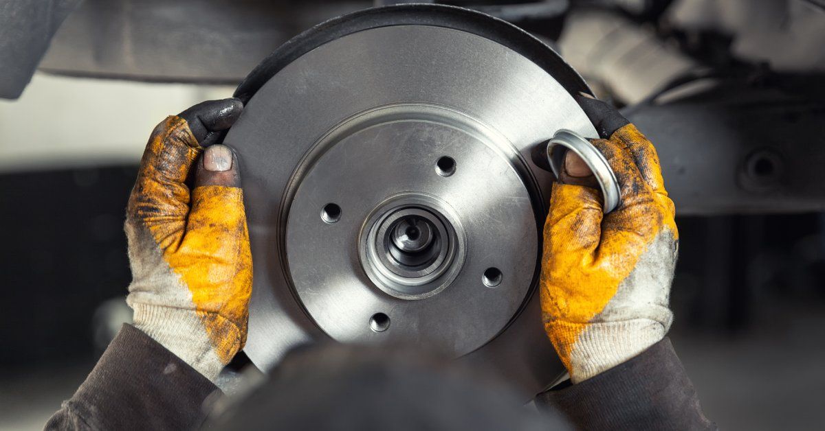 A person wearing yellow and white fingerless gloves is installing a clean new metal brake rotor on a vehicle.