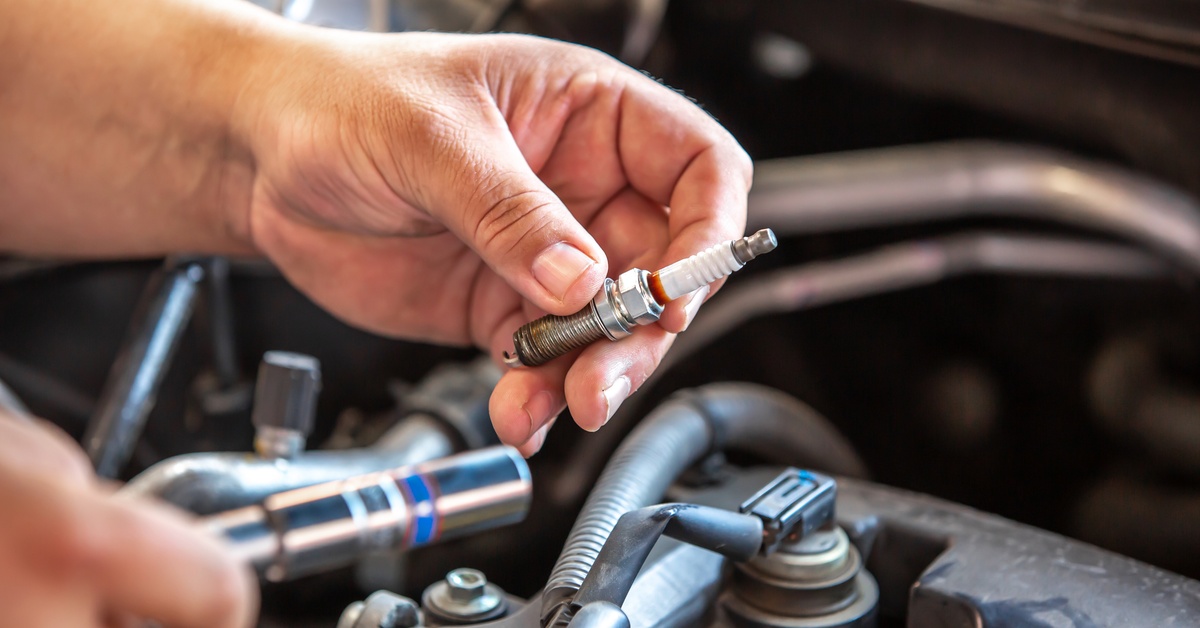 A person is holding a used spark plug in their hands. They are holding the spark plug over a car engine. A person is holding a used spark plug in their hands. They are holding the spark plug over a car engine.