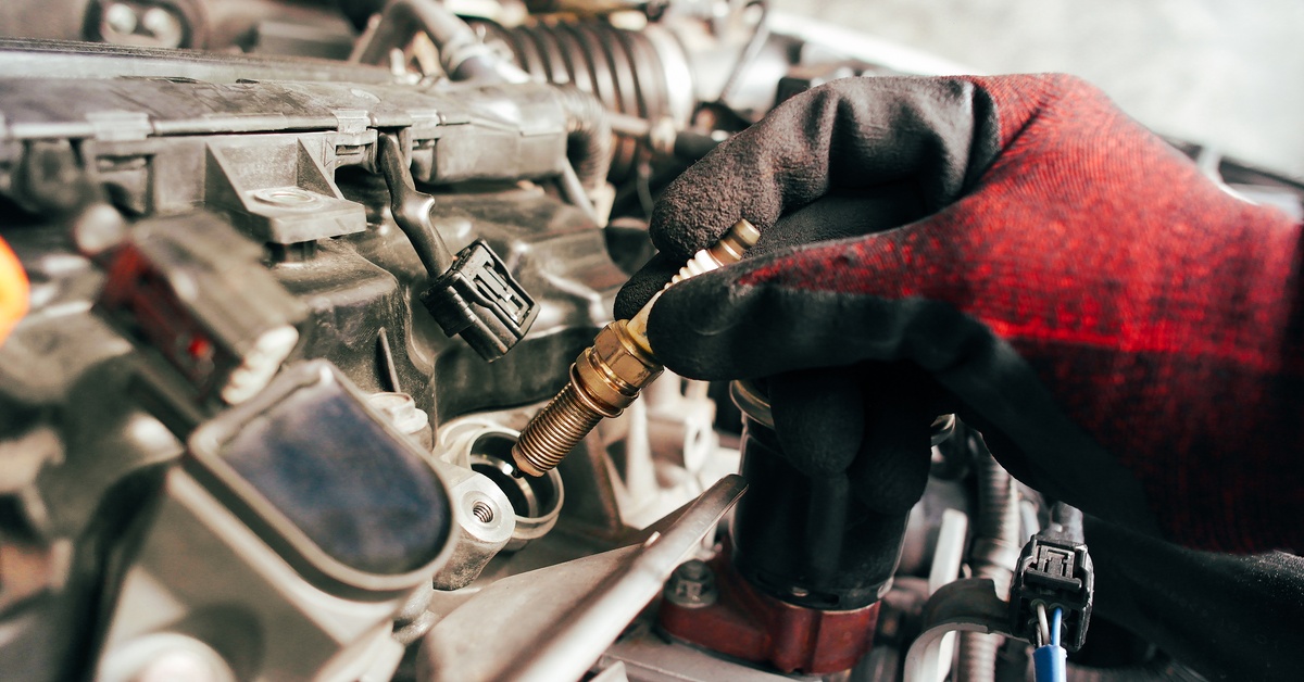 A person wearing red-and-black gloves is inserting a brand-new spark plug into the engine of a car. A person wearing red-and-black gloves is inserting a brand-new spark plug into the engine of a car.