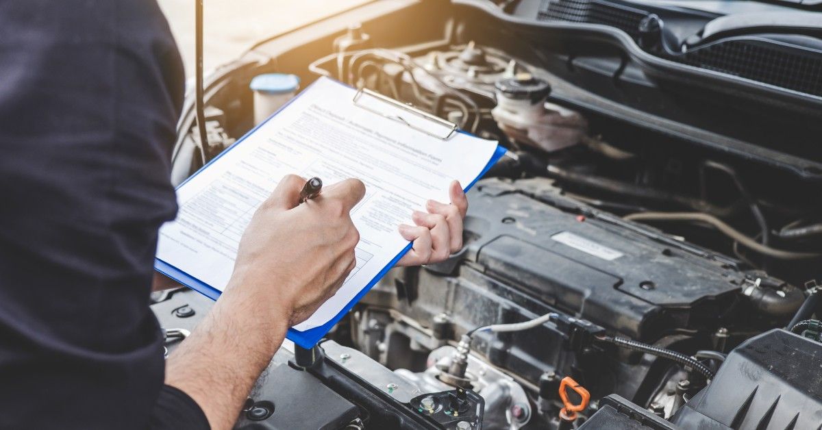 A person is standing over a car engine. They are holding a pen and a blue clipboard with documents on it. A person is standing over a car engine. They are holding a pen and a blue clipboard with documents on it.