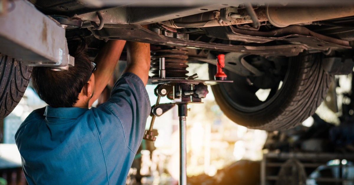 A mechanic is working on the underside of a car in an auto shop. A jack is underneath the vehicle for support. A mechanic is working on the underside of a car in an auto shop. A jack is underneath the vehicle for support.