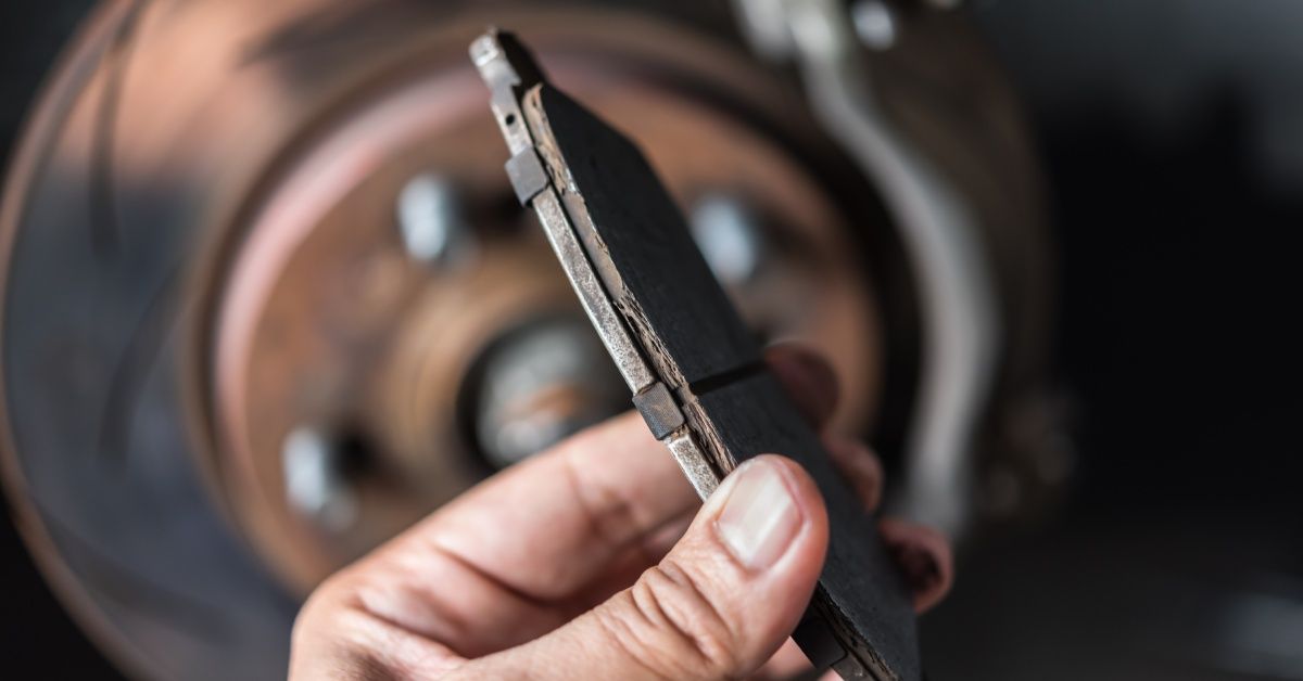 A person is holding a brake pad in their bare hands in front of a tire. The brake pad is visibly deteriorating.